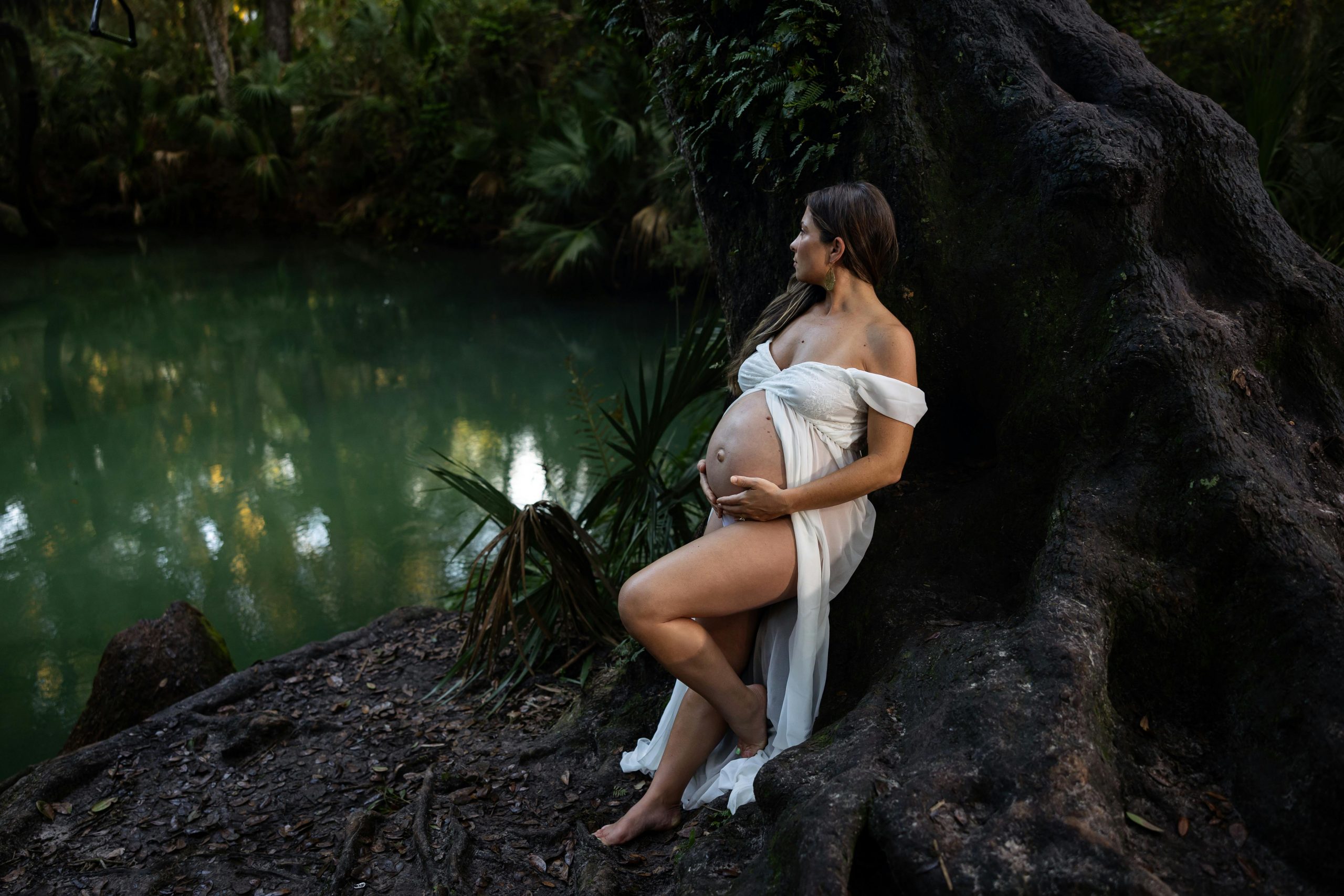 pregnant woman cradling her bump from underneath while leaning against a tree by a river