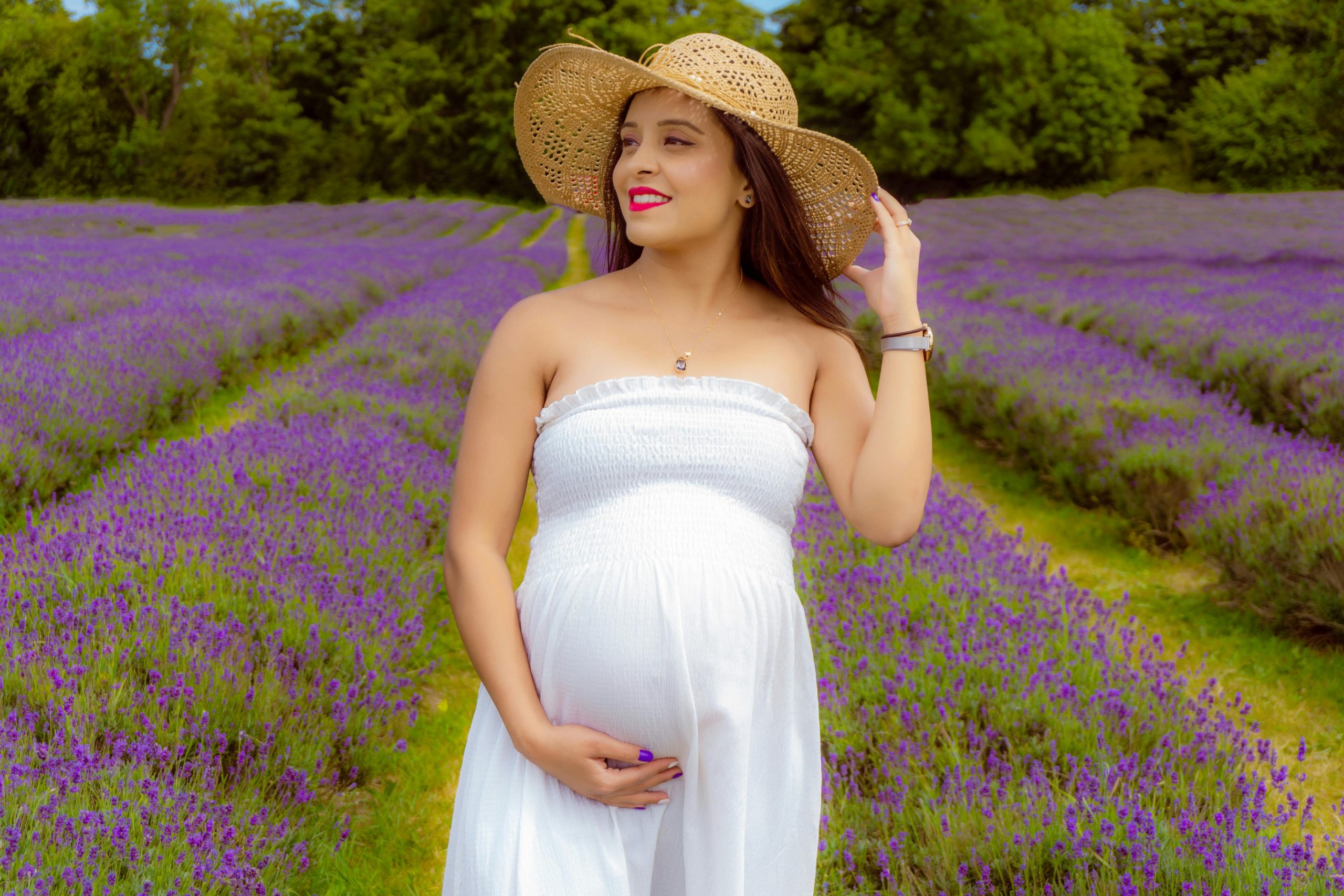 pregnant woman in a white dress and wide-brimmed hat, posing with a flower field behind her